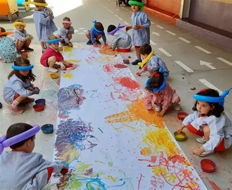 Group of young children in school uniforms and colorful hats painting together on a large white canvas on the floor with vibrant paint colors