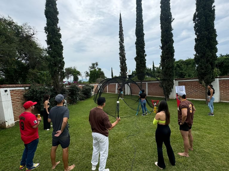 Group of people gathered in a grassy outdoor area with tall cypress trees and brick buildings in the background