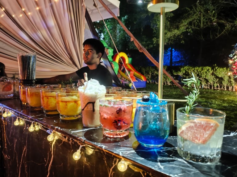 Outdoor bar setup at night with colorful cocktails on a decorated table, bartender in background, string lights and tropical foliage visible