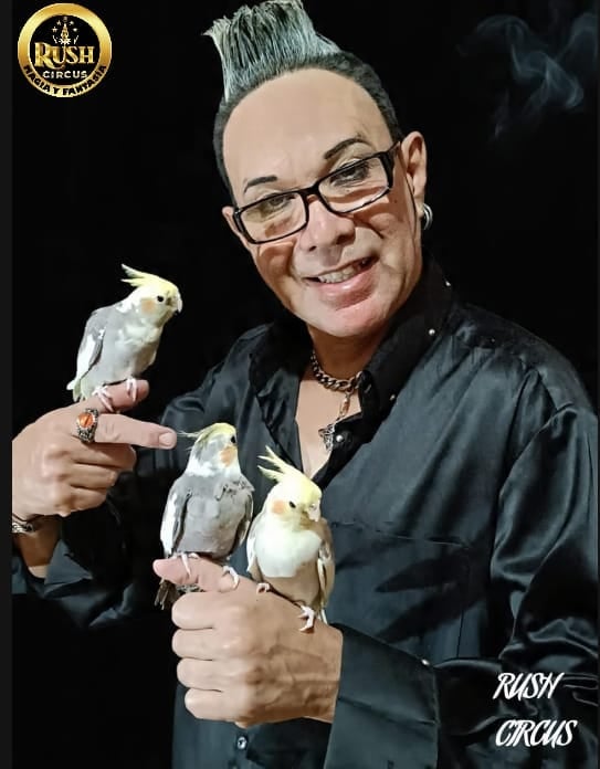 Man with spiky gray hair and glasses holding three white cockatiel birds, wearing black shirt with Rush Circus logo