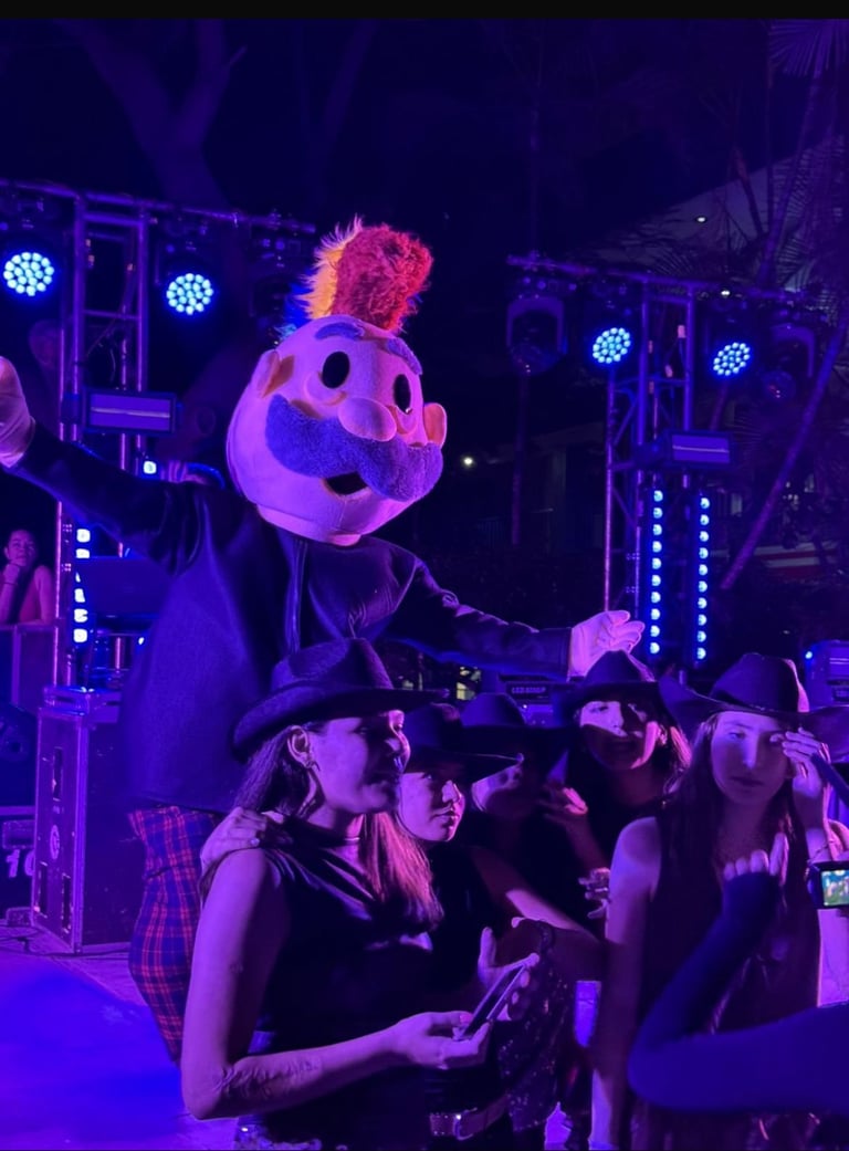 Group of people at a nighttime concert or event surrounded by purple and blue stage lighting, with a giant glowing pumpkin-shaped balloon with a jack-o-lantern face above them.
