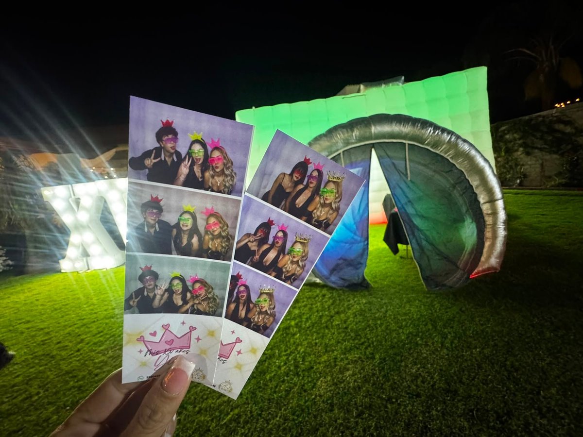 Person holding photo booth strips at night with illuminated green inflatable structure in background on grass