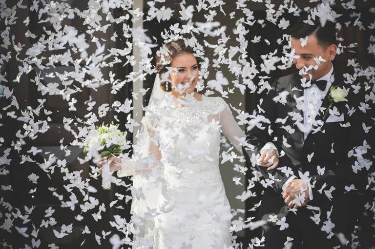 Bride and groom in formal attire under a floral backdrop with white petals falling during their wedding celebration